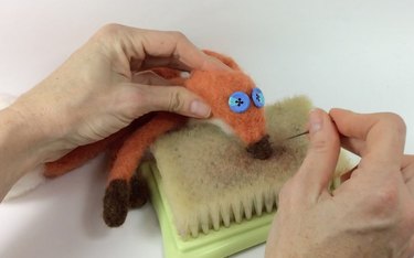 Female hands needle felting a brown nose onto a fox face.