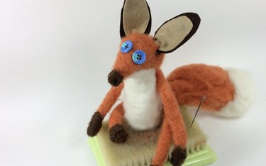 Needle felted fox on felting pad next to felting needle.