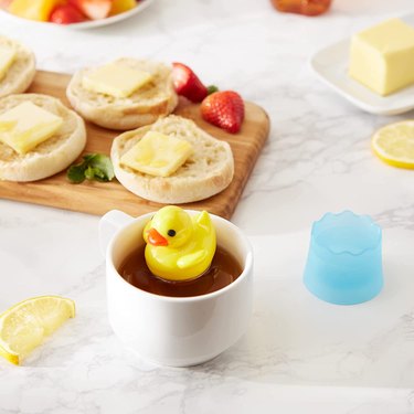 Rubber ducky tea infuser, floating in a white mug on a marble countertop with buttered English muffins and berries