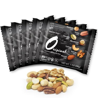 Individual snack packs of Roastery Coast mixed nuts in their black packaging, fanned out on a white ground above a handful of the shelled mixed nuts
