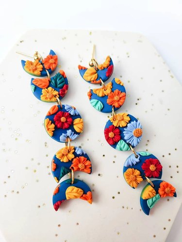 Dangly earrings shaped like various phases of the moon featuring red, orange and blue flowers on a blue background