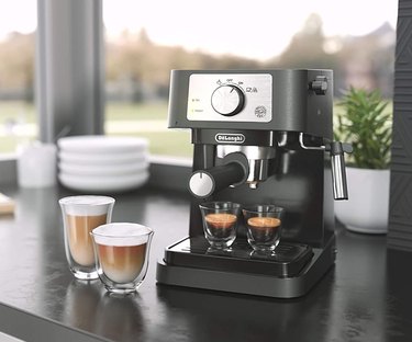 Silver and black manual espresso machine next to two cups of cappuccinos on countertop.