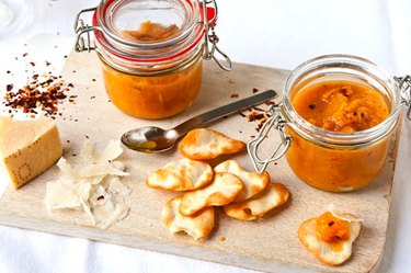 Jars of English peach and ginger chutney on wooden board with Parmesan and chips.