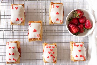 Strawberry jam tarts with playing card decorations on a wire rack, next to a bowl of strawberries