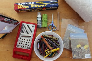 An iron, cheese grater, broken crayons, acrylic picture frames, wax paper, paper towels, pencil, pair of scissors, spool of string, and bottle of craft paint