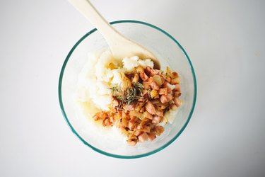 Spoon combining the onion and bacon with the potato in a glass bowl.