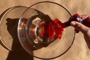 A hand dripping red food coloring into a clear bowl filled with water and corn syrup