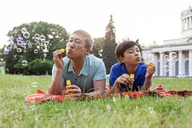 asian old father together with son lie on the grass in the park and blow soap bubbles and rejoice in summer