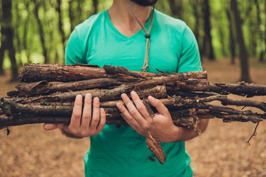 Cropped close up photo of a bearded male tourist during hiking, holding wood for a campfire, in green t shirt, in a forest in the early fall