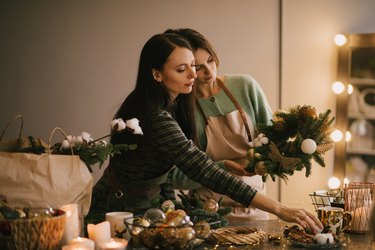 Two millennial women making Christmas wreath using pine branches and festive decorations. Small business