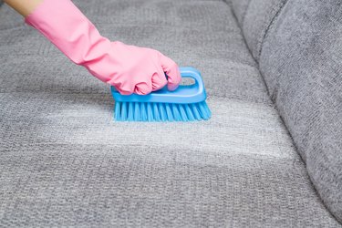 Dry cleaner's employee hand in protective gloves removing stain from gray sofa with brush and special foam. Early spring regular cleanup. Commercial cleaning company. Close up. Side view.