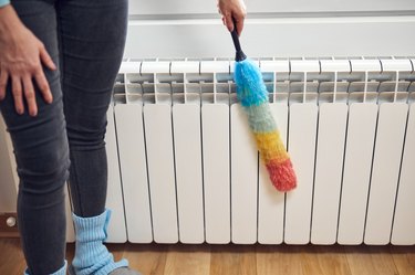 Woman with a dust stick cleaning central heating gas radiator at home.