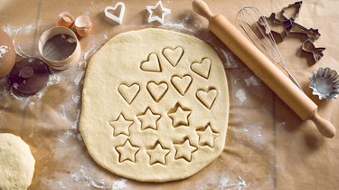 Cookie dough with cutters and cut cookies in various shapes, shown on a countertop with rolling pin, utensils and baking ingredients