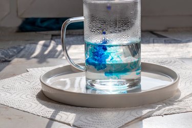 Glass mug with hot water, into which blue food coloring is dripped.