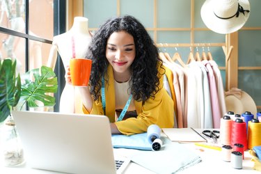 Young women fashion designer using laptop working and design tailor and cloth in the showroom, Lifestyle Stylish tailor taking measurements on mannequin for cutting cloths in studio.