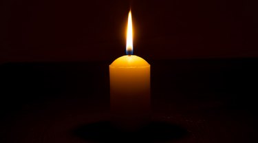 Single burning candle flame or light glowing on a big white candle on black or dark background on table in church for Christmas, funeral or memorial service with copy space.